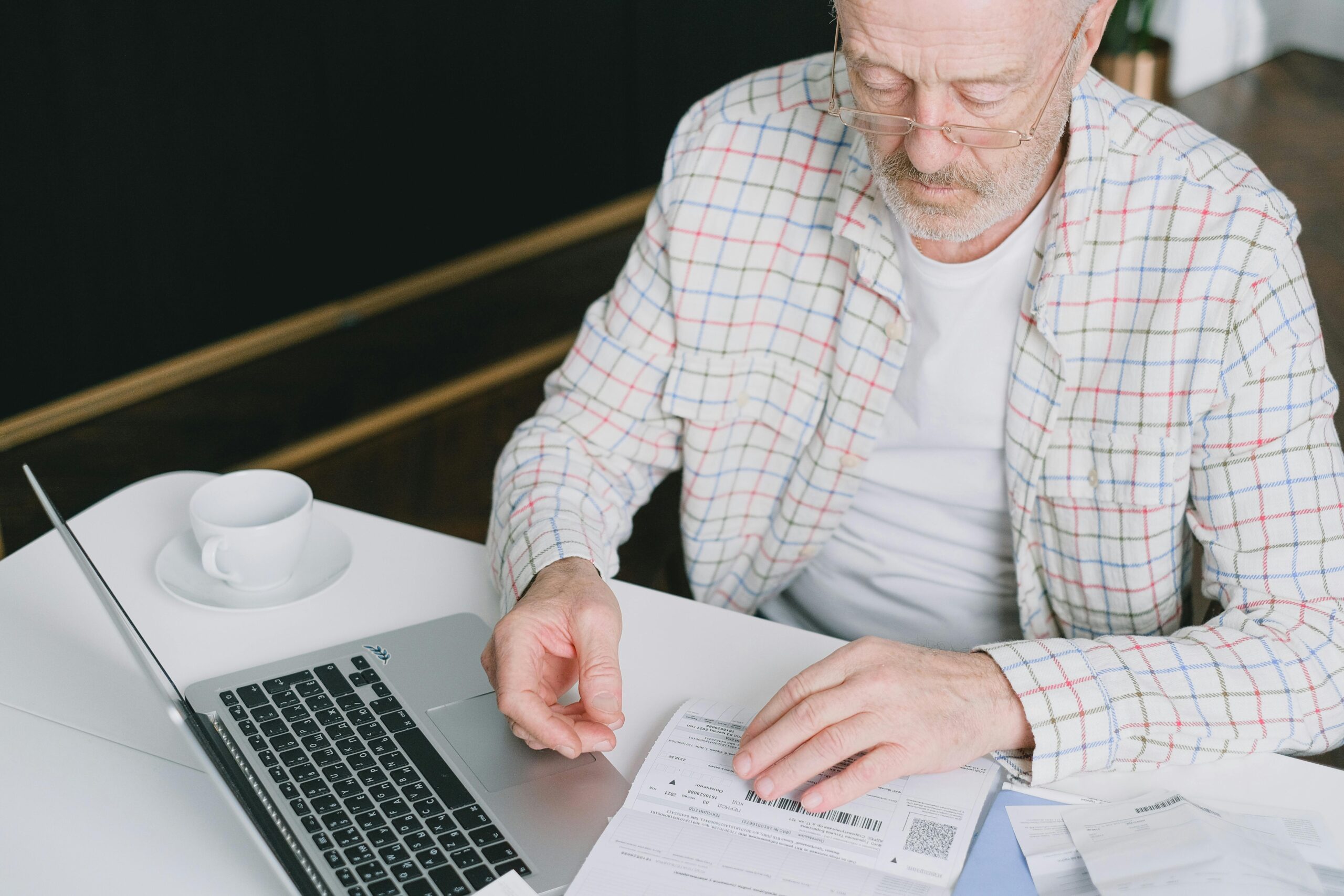 Elderly man reviewing financial documents at a table with a laptop